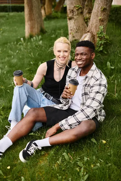 Una pareja feliz y multicultural, un hombre afroamericano y una mujer caucásica, sentados juntos en el césped en un parque. - foto de stock