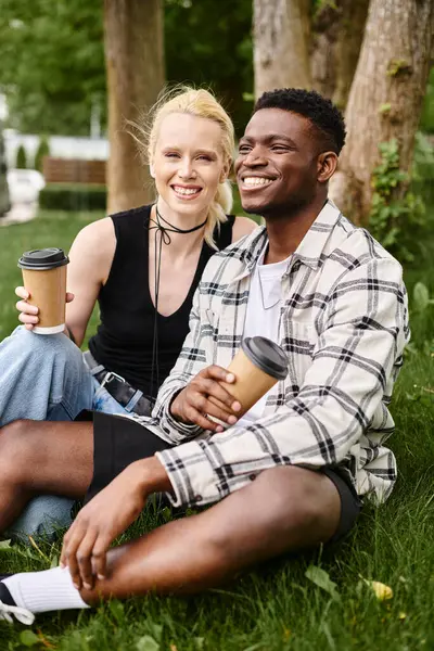 Una pareja multicultural, un hombre afroamericano y una mujer caucásica, se sientan juntos en la exuberante hierba verde de un parque. - foto de stock