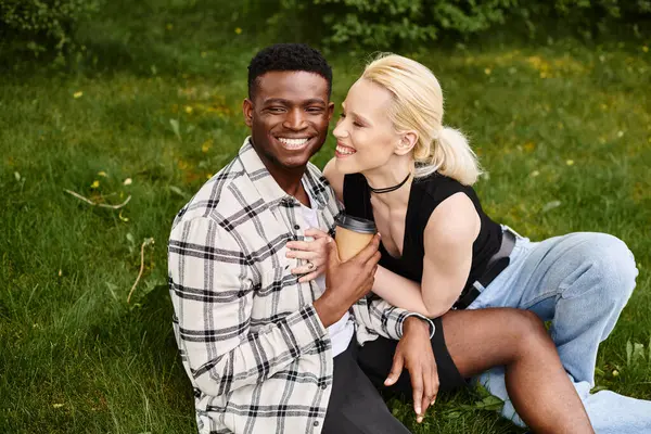 Um homem afro-americano feliz e uma mulher caucasiana sentam-se juntos na grama, apreciando a beleza do ar livre. — Fotografia de Stock