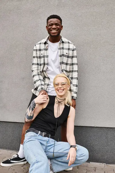 A man and woman of different ethnicities sitting closely together on the ground next to a grey building in an urban setting. — Stock Photo