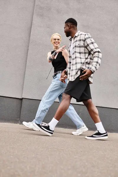 Una feliz pareja multicultural camina por una calle urbana junto a un edificio gris, disfrutando de la compañía de los demás. - foto de stock