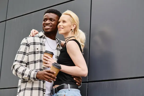 A happy, multicultural couple standing next to each other on an urban street near a grey building. — Stock Photo