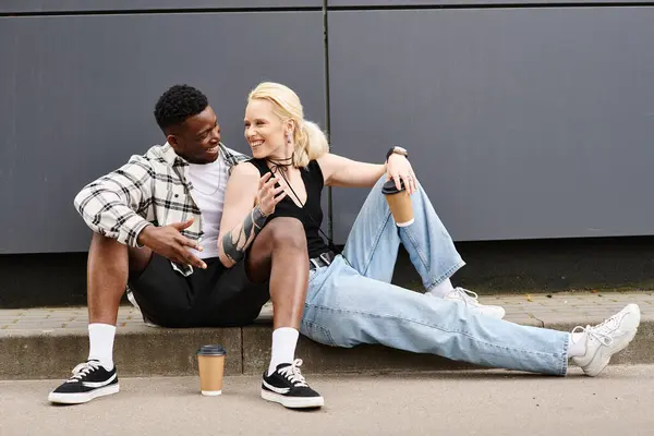 Ein Mann und eine Frau verschiedener Ethnien sehen zufrieden aus, als sie nebeneinander auf dem Boden in der Nähe eines grauen Gebäudes in der Stadt sitzen.. — Stockfoto