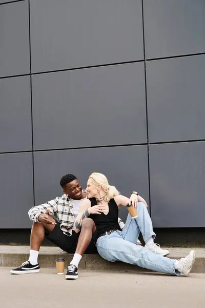 Щаслива мультикультурна пара сидить поруч один з одним на землі біля сірої міської будівлі, ділячи спокійний момент. — стокове фото