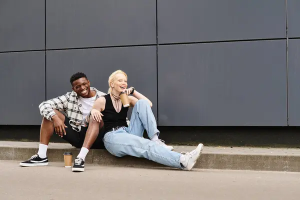 Ein fröhliches multikulturelles Paar sitzt nebeneinander auf dem Boden in der Nähe eines grauen städtischen Gebäudes — Stockfoto