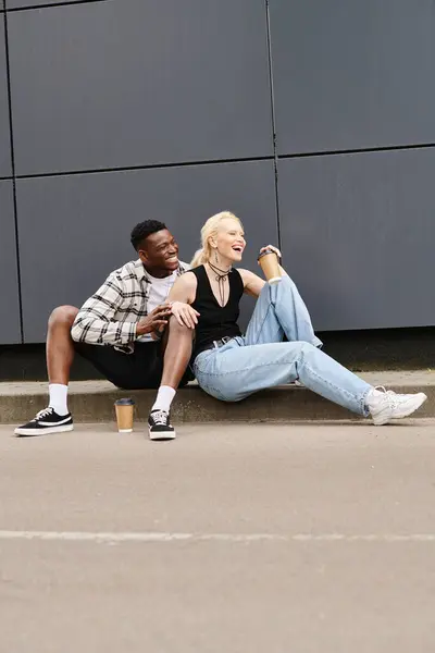 A happy multicultural man and woman sitting together on the ground near a grey building, enjoying their time outdoors. — Stock Photo