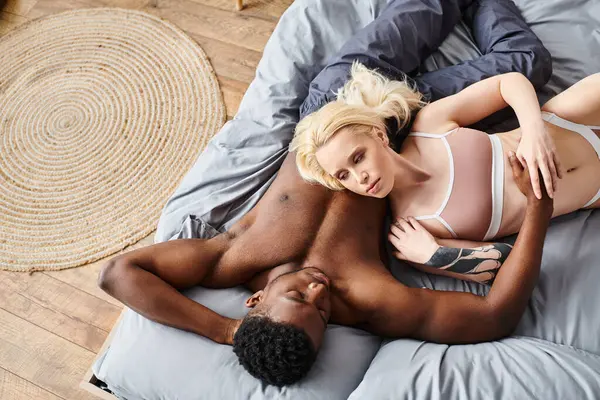 Un novio y una novia multiculturales están tendidos románticamente entrelazados en una cama en casa, compartiendo un momento de intimidad juntos. - foto de stock