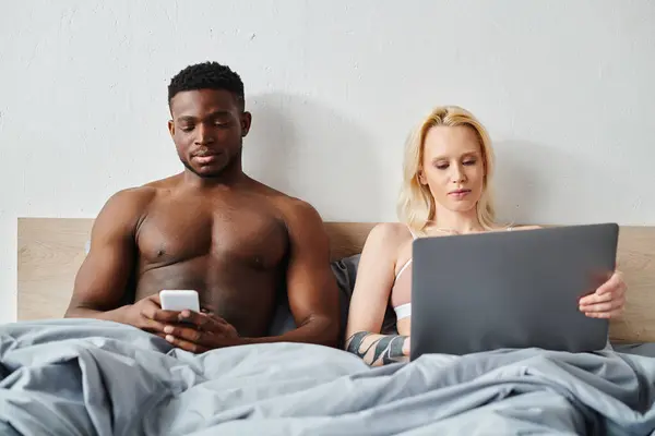 A multicultural boyfriend and girlfriend sit on a bed, focused on their devices. — Stock Photo