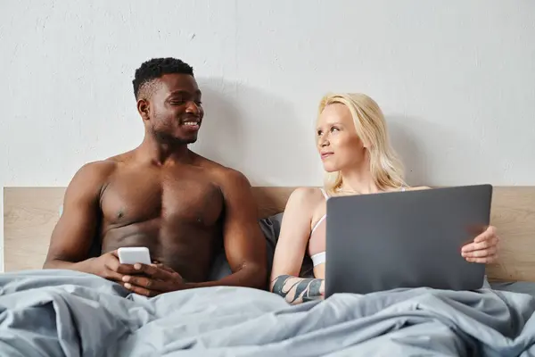 A multicultural man and woman sit closely on a bed, engrossed in the screen of a laptop before them. — Stock Photo
