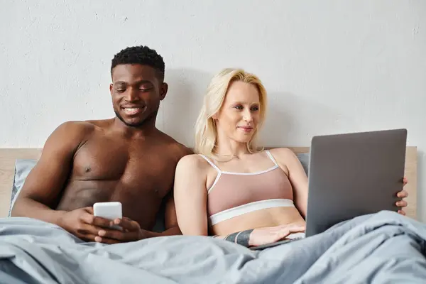 A multicultural couple sits on a bed, engrossed in a laptop screen together. — Stock Photo