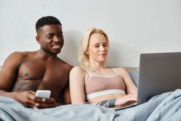 Multicultural couple sitting on bed, engrossed in laptop screen. — Stock Photo