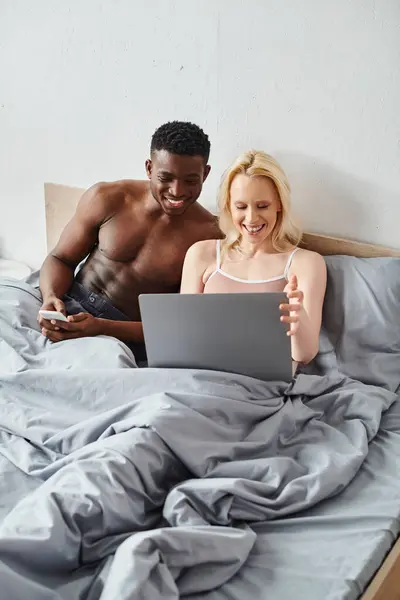 Multicultural boyfriend and girlfriend sitting on bed, engrossed in a laptop screen. — Stock Photo
