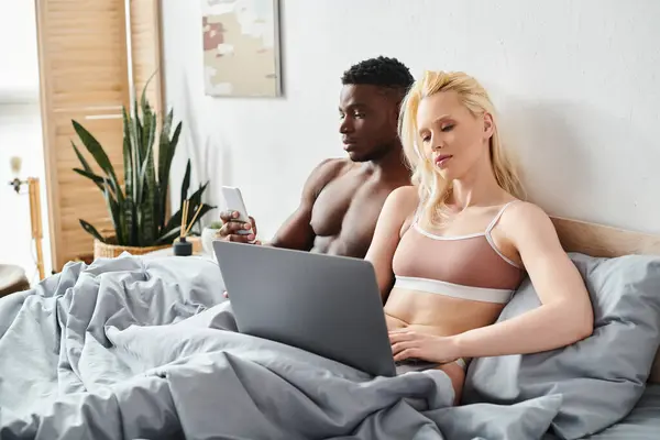 Un hombre y una mujer multiculturales están sentados en una cama, absortos en la pantalla de un ordenador portátil en un entorno íntimo y acogedor. - foto de stock