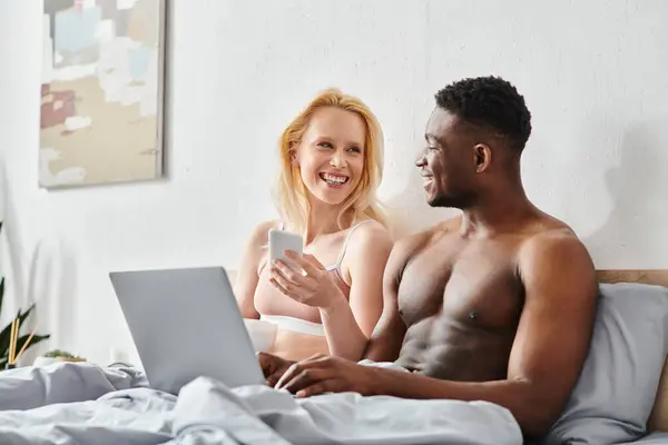 Una coppia multiculturale, un uomo e una donna, si siedono insieme su un letto, avvolti nello schermo di un computer portatile. — Foto stock