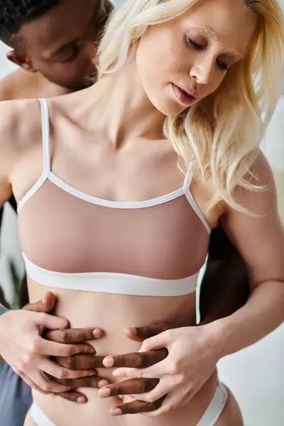Una mujer multicultural acostada en la cama con su pareja, sosteniendo tiernamente su estómago en un sujetador blanco. - foto de stock