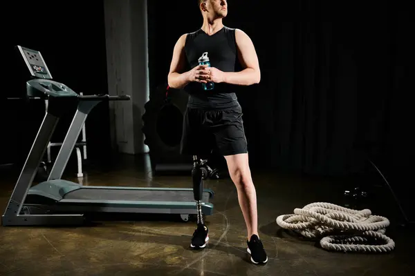 Un handicapé avec une prothèse de jambe se tient devant un tapis roulant, tenant une bouteille d'eau dans un cadre de gymnase. — Photo de stock