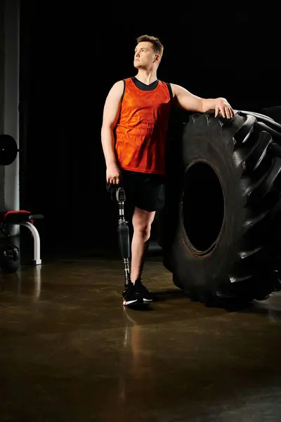 Um homem com uma perna protética ao lado de um pneu enorme num ginásio. — Fotografia de Stock