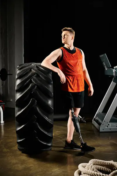 Un uomo disabile con una gamba protesica si erge orgogliosamente accanto a un grande pneumatico in palestra, mostrando la sua determinazione e forza.. — Foto stock