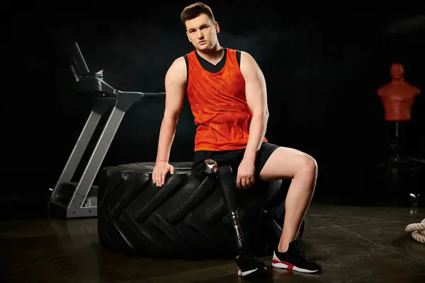 Un hombre discapacitado con una pierna protésica se sienta con confianza sobre un neumático negro, haciendo ejercicio en un gimnasio. - foto de stock