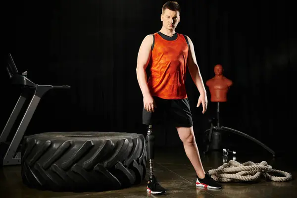 Un hombre con una pierna protésica en una camisa naranja está de pie junto a un neumático. - foto de stock