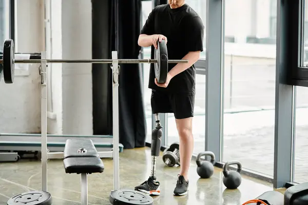 Un hombre discapacitado con una pierna protésica se para en un gimnasio, sosteniendo un bar mientras se ejercita para construir fuerza y resistencia. - foto de stock