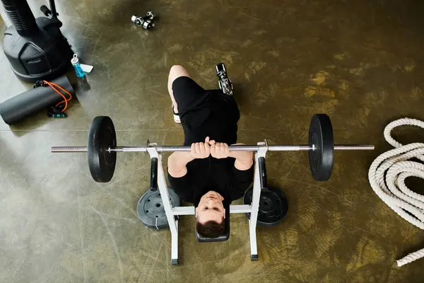 Una persona con una pierna protésica está en un banco, levantando una barra en el gimnasio. - foto de stock