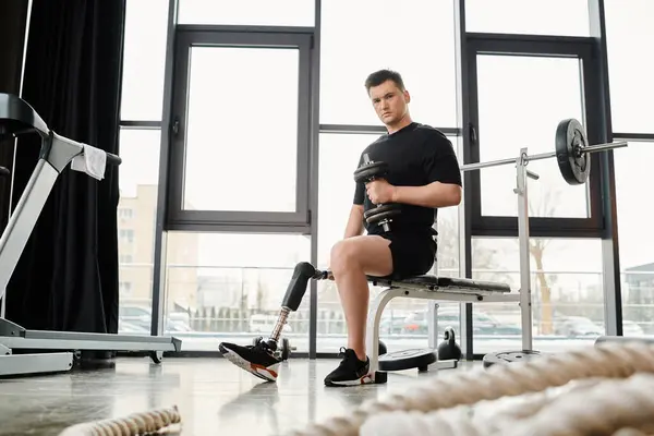 Un homme avec une prothèse de jambe est vu assis sur un banc dans une salle de gym, travaillant avec haltère — Photo de stock