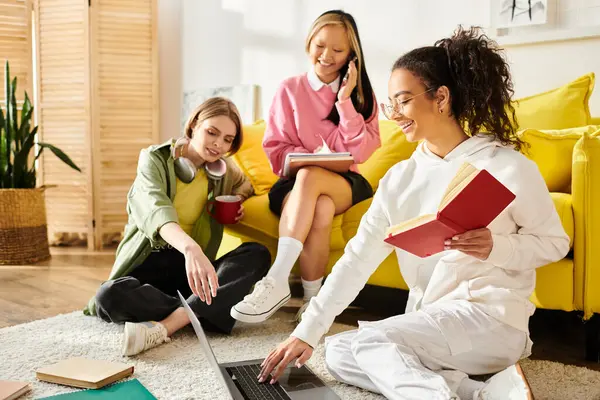 A multicultural group of young women happily sit together on a bright yellow couch, sharing laughter and studying. — Stock Photo