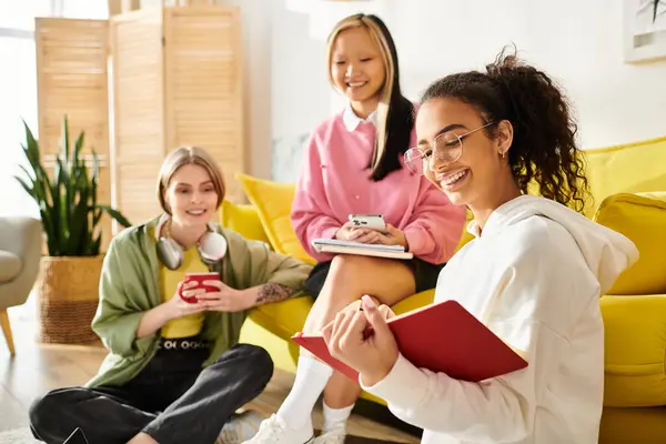 Diverse Teenager-Mädchen sitzen gemütlich auf einer leuchtend gelben Couch und studieren gemeinsam von zu Hause aus. — Stockfoto