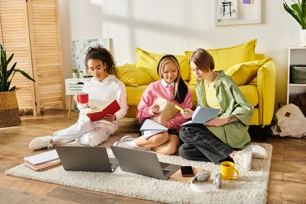 Tres adolescentes de diferentes razas están sentadas en el suelo, absortas en sus computadoras portátiles, trabajando en un proyecto juntas. - foto de stock
