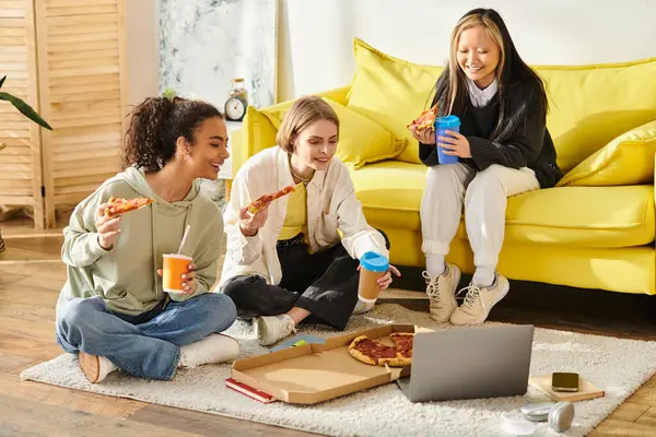 Tres adolescentes de diferentes razas se sientan en el suelo, disfrutando de pizza y café juntas en un ambiente acogedor. - foto de stock