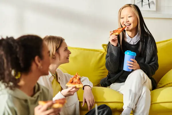 Tres adolescentes de diferentes razas se sientan en un sofá amarillo, disfrutando de la pizza juntas. - foto de stock