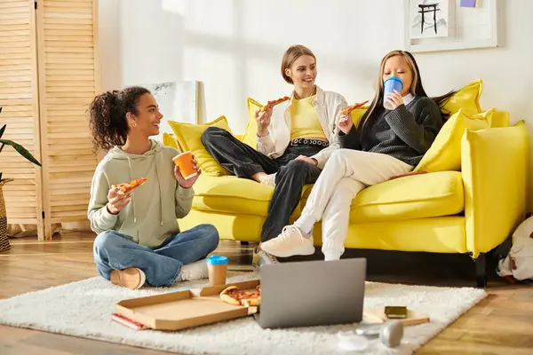 Tres adolescentes diversas se sientan en un sofá amarillo, se unen sobre la pizza y la risa. - foto de stock