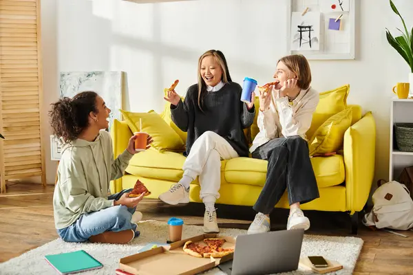 Tres adolescentes, de diferentes etnias, están sentadas en un sofá amarillo brillante, disfrutando rebanadas de pizza juntas. - foto de stock