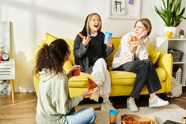 Tres adolescentes de diferentes razas felizmente se sientan en un sofá amarillo brillante, charlando y comiendo rebanadas de pizza. - foto de stock