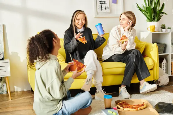 Tres chicas adolescentes disfrutan de la pizza en un sofá amarillo en un ambiente acogedor en casa. - foto de stock