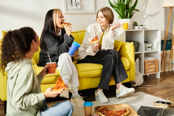 Tres adolescentes interracial se sientan alegremente en un sofá amarillo, pegándose a la pizza. - foto de stock