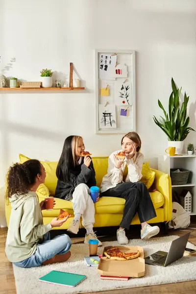 Tres mujeres, de diferentes etnias, disfrutan de rebanadas de pizza mientras están sentadas en un sofá amarillo brillante. - foto de stock
