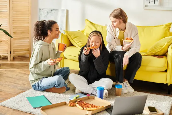Троє молодих жінок різних рас і стилів сидять на підлозі, насолоджуючись піцою разом у затишній обстановці. — стокове фото