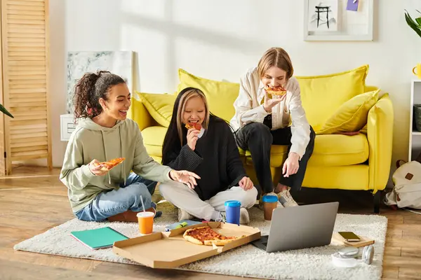 Tres mujeres jóvenes, que representan la diversidad, se sientan en el suelo disfrutando de rebanadas de pizza juntas en un acogedor entorno hogareño. - foto de stock
