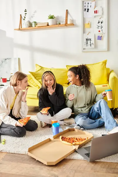 Adolescentes, de diferentes razas, se sientan en el suelo comiendo felices rebanadas de pizza juntas en una acogedora reunión. - foto de stock