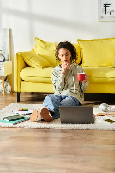 A young woman sits on the floor, gracefully clutching a warm cup of coffee while studying on her laptop at home. — Stock Photo