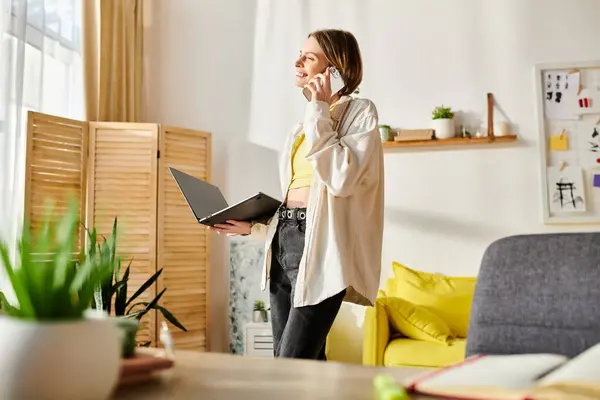 Eine junge Frau in einem modernen Wohnzimmer führt im Stehen ein Telefongespräch, den Laptop vor sich offen. — Stockfoto