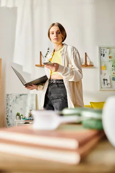 Una giovane donna in una stanza, immersa nello studio mentre in piedi con un computer portatile nelle vicinanze. — Foto stock