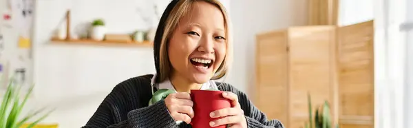 A young woman, enjoying a moment of laughter, holds a red cup in her hand while studying at home — Stock Photo