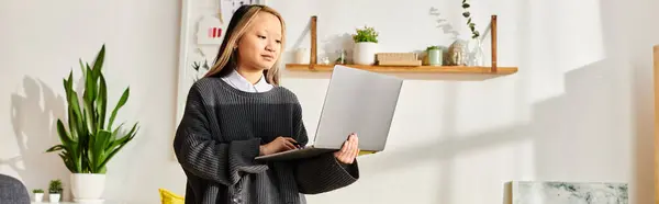 A young Asian girl immersed in digital learning, standing in a living room while holding a laptop. — Stock Photo