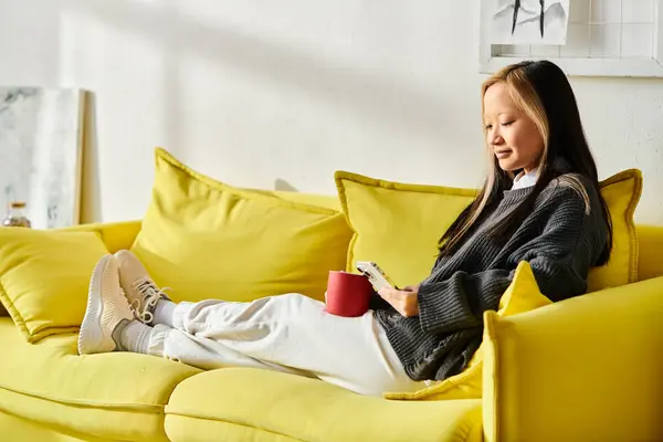 A young Asian woman sits peacefully on a yellow couch, holding a red cup and using smartphone — Stock Photo