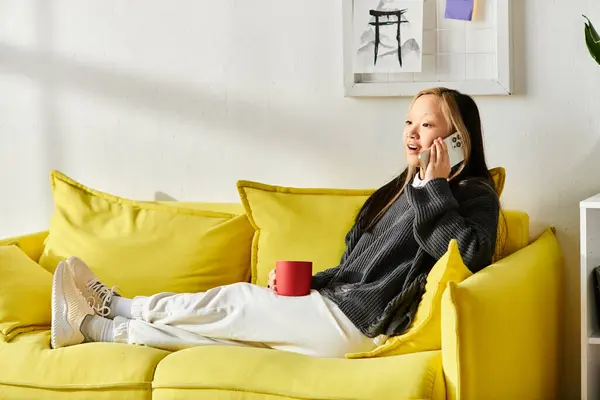 Una joven asiática está cómodamente sentada en un sofá amarillo, participando en una animada conversación en su teléfono celular. - foto de stock