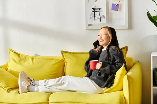 Una joven asiática está sentada en un sofá amarillo, sosteniendo una taza de café en casa. - foto de stock