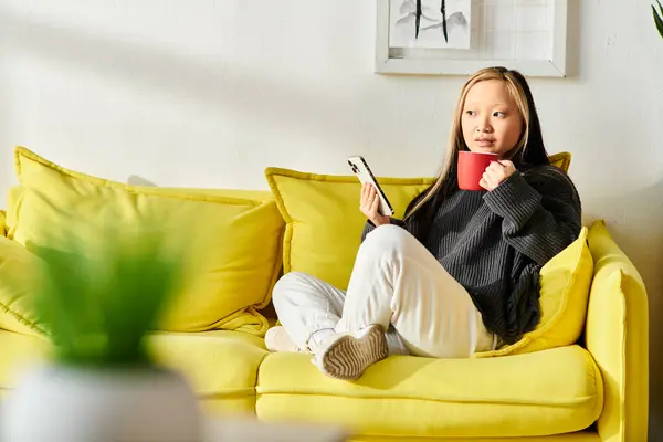 A young woman of Asian descent sits on a yellow couch, holding a cup of coffee in her hands while taking a break from e-learning with her smartphone. — Stock Photo